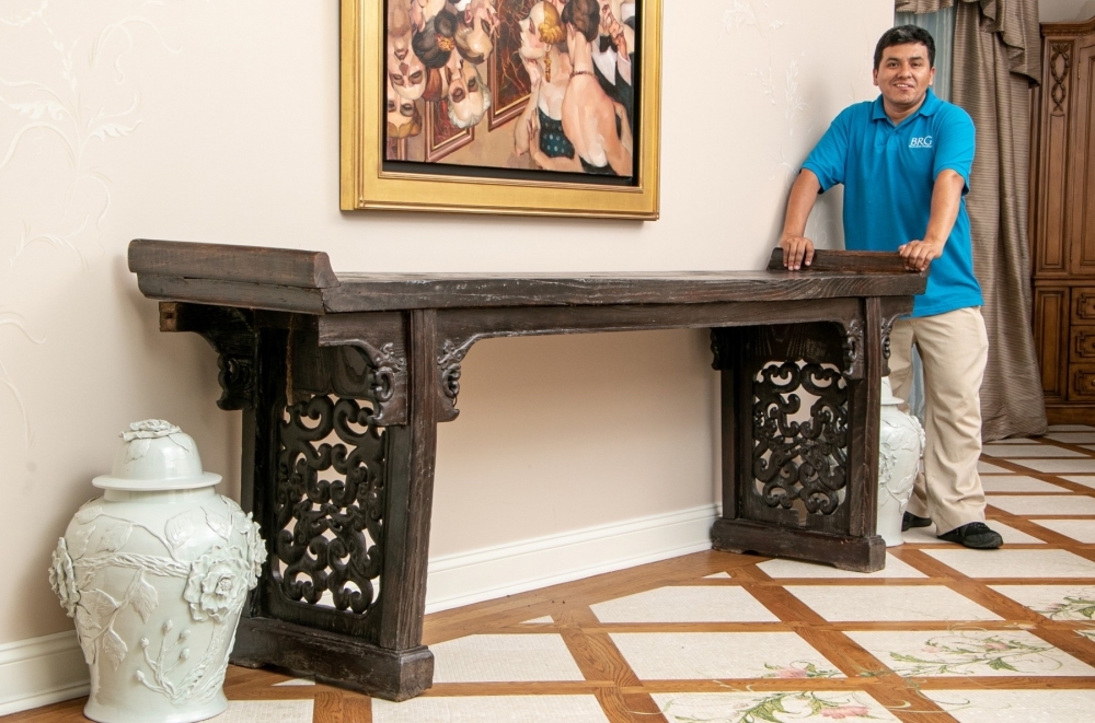 A long elmwood Ming Dynasty altar table with intricate carved inset side panels, planked top, carved corner brackets and winged table top ends. A pair of ivory-toned tall covered ginger jars with raised floral embellishments sits on either side of the table on the floor and an employee with a turquoise blue shirt and tan pants stands on the right side of the table.